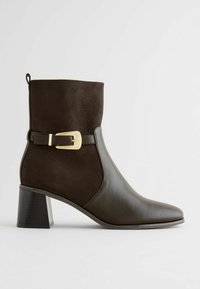 Bottine marron avec une tige en suede, une partie inférieure en cuir, un bout pointu et un accent de boucle dorée au niveau de la cheville. Dispose d'un talon carré.