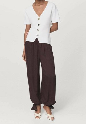 Femme portant un haut blanc à manches courtes avec de gros boutons, un pantalon ample marron foncé cintré aux chevilles par des nœuds, et des sandales blanches à talons.