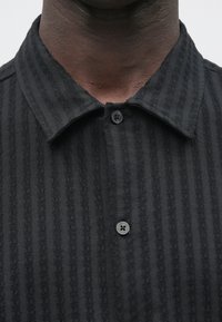 Zwart overhemd met kraag van gestructureerde stof met verticale strepen, voorzien van een knoopsluiting en twee zwarte knopen.