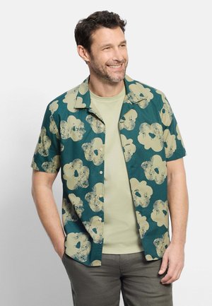 Homme souriant portant une chemise verte à manches courtes ouverte avec de grandes impressions florales beiges sur un t-shirt vert clair et un pantalon gris.