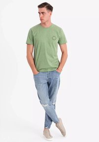 Grønprikket t-shirt med rund halsudskæring, parret med lyseblå slidte jeans og beige sneaker. Modellen står afslappet med hænderne i lommen.