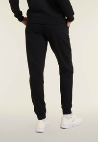 Persona che indossa pantaloni jogger neri, calzini bianchi e scarpe sportive bianche, in piedi con le mani in tasca contro uno sfondo neutro.