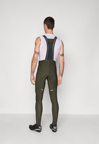 Collants de vélo en tissu rembourré vert olive foncé, avec des bretelles en mesh noir et des accents réfléchissants blancs sur les jambes.