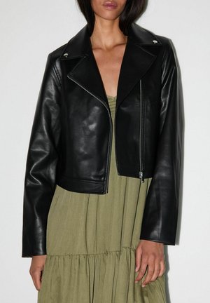 Vrouw draagt een zwarte cropped leren jas over een olijfgroene geplooide jurk met lange mouwen en reverskraag.
