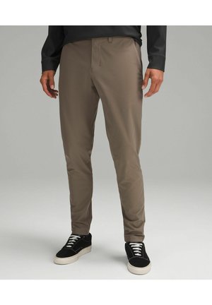 Homme portant un pantalon marron ajusté et des baskets noires avec des semelles et des lacets blancs, debout devant un fond gris uni.