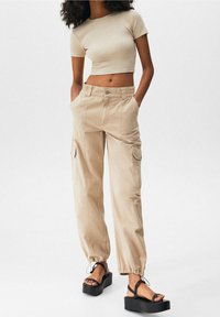 Pantalon cargo beige avec poignets élastiques et multiples poches, associé à un court haut beige et des sandales à plateforme noires. Texture lisse.