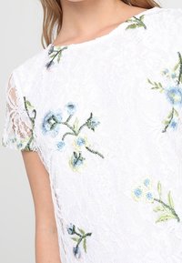 White lace top featuring floral embroidery in blue and green, with short sleeves and a round neckline. Delicate texture and layered design.