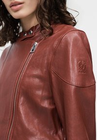 Γυναίκα με καφέ δερμάτινο μπουφάν με ασύμμετρο ασημένιο φερμουάρ και ανάγλυφο λουλουδένιο λογότυπο στον ώμο, με εμφανή σγουρά μαλλιά.