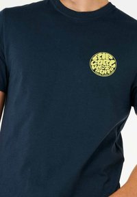 Mørkeblå bomulds t-shirt med korte ærmer. Har et rundt gult grafisk logo på venstre brystområde med tekst og bølget design.