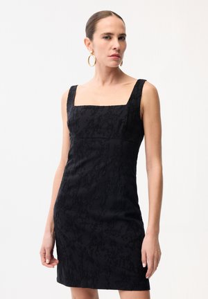 Vrouw die een mouwloze zwarte jurk met textuur en een vierkante halslijn draagt, met gouden oorringen, staand tegen een witte achtergrond.