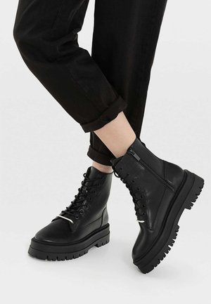 Černé kožené kotníkové boty s robustní gumovou podrážkou, šněrováním vpředu a bočním zipem. Mají hladký povrch a pevný design.