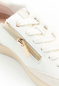 Sneaker bianca con suola crema, lacci bianchi e un dettagli di cerniera dorata lucida vicino all'area dei lacci.