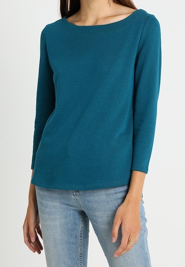 Haut à manches longues couleur sarcelle avec un col bateau et une texture douce, associé à un jean bleu clair. Présente une coupe décontractée et un ourlet cousu.