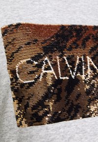 Szary sweatshirt z prostokątną łatą cekinów w kolorze brązowym, czarnym i złotym, z napisem "CALVIN" w białym kolorze.