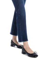 Zapatos de charol negro con tacón cuadrado y punta redondeada, con un detalle de lazo en la parte frontal, combinados con unos vaqueros azules recortados y deshilachados.