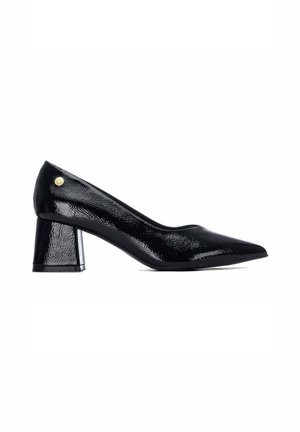 Zapato de tacón para mujer de charol negro con tacón de bloque redondeado y un pequeño logo circular dorado cerca del talón.
