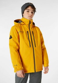 Giacca impermeabile gialla con cappuccio, dotata di zip e dettagli neri, polsini regolabili e due tasche sul petto. Abbinata a un berretto grigio.