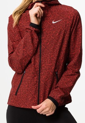 Frau trägt eine rote, gemusterte Nike Jacke mit Reißverschluss und Kapuze, sie richtet den Reißverschluss an ihrem Hals.