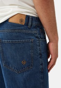 Mörkblå denimjeans med en slät yta. Har en beige läderlapp vid midjan och en diamantlogotyp broderad på bakfickan.