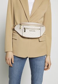 Sac banane Michael Kors blanc et beige avec un motif à logo MK signature, deux poches zippées et des accessoires en or, porté sur un blazer beige.