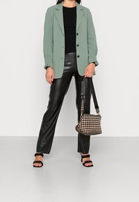 Blazer verde con rever a punta, top nero a costine, pantaloni neri in ecopelle, tacchi strappy neri e una borsa a scacchi marrone.