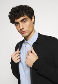 Giacca nera zip-up sopra una camicia button-up azzurro chiaro. La giacca ha una texture liscia e un colletto; dettagli dei bottoni visibili sulla camicia.