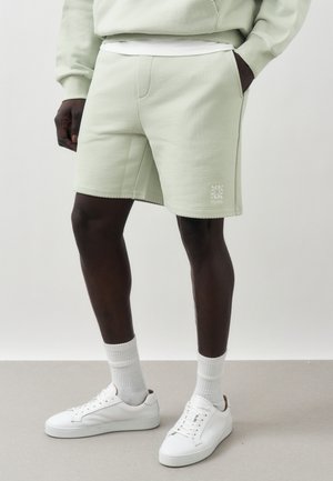 Mann iført lys grønne shorts, hvite sokker, hvite joggesko og en matchende genser, står med en hånd i lommen.