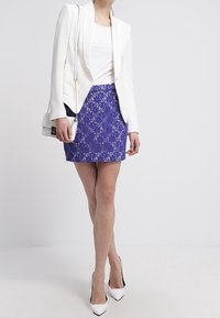 Blazer blanco sobre una blusa blanca ajustada, combinado con una falda de encaje azul y tacones blancos de punta. Accesorios con un pequeño bolso bandolera.