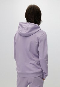 Felpa lilla con cappuccio regolabile, morbido al tatto, con polsini e bordo a coste, e vestibilità ampia, indossata con pantaloni coordinati.