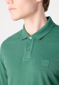 Polo verde din material moale, cu guler clasic, două butoane și un patch cu logo ton pe pieptul stâng.