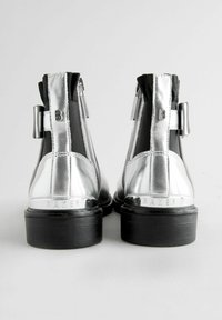 Botas de tobillo plateadas con un acabado metálico elegante, paneles elásticos laterales y acentos negros. Cuentan con cierres de cremallera y una suela negra plana.