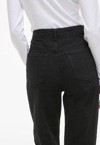 Crne traperice visokog struka s opuštenim krojem, koje imaju dva strašnja džepa i glatku teksturu denima. Nosive s bijelom dugom majicom.