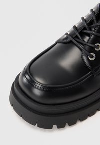 Pantofii din piele neagră, cu șireturi, au o talpă din cauciuc groasă și adâncită, cu găici metalice, prezentați din unghiul de sus-frontal pe un fundal neutru.