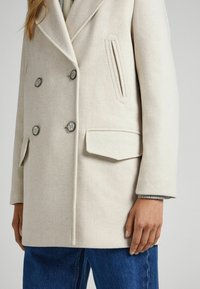 Double-breasted beige coat made of textured fabric, featuring large buttons, side pockets, and a slim fit with a notch lapel design.