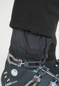 Pantalons de ski noirs retroussés sur une chaussure de ski noire, avec une boucle métallique et des accents texturés. La chaussure dispose d'un cadran rond pour les réglages.