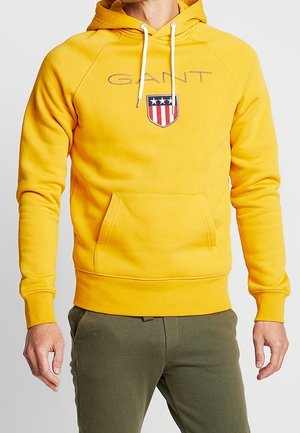 Man wearing yellow GANT hoodie with white drawstrings and shield logo, paired with olive green pants, standing with arms relaxed.