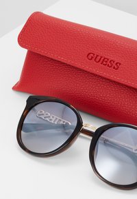 Occhiali da sole tartarugati con lenti sfumate, abbinati a una custodia in pelle rossa testurizzata impressa con la scritta "GUESS." Forma tondeggiante, dettagli dorati.