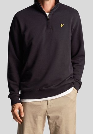 Mann trägt einen schwarzen Pullover mit Viertel-Reißverschluss und gelbem Vogel-Logo, weißes Unterhemd und beigefarbene Hose, steht mit einer Hand in der Tasche.