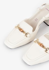 Zapatos de piel blanca tipo slingback con puntera cuadrada, herrajes dorados y un accesorio decorativo con detalle de carey. Tacón bajo y cuadrado.