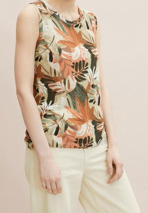 Top bez rękawów z wielokolorowym tropikalnym nadrukiem liści w odcieniach brązu, zieleni i bieli, noszony z jasnobeżowymi spodniami na stojącej osobie.