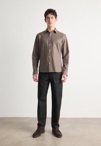 Chemise marron à manches longues avec un design boutonné, associée à un pantalon en denim foncé et des chaussures marron, debout devant un fond neutre.