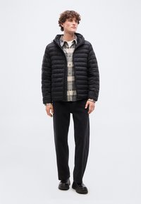 Νέος άνδρας που φοράει μαύρο πουπουλένιο μπουφάν πάνω από μπεζ πουκαμίσα με καρό, μαύρο παντελόνι και μαύρα παπούτσια, στέκεται με ουδέτερη έκφραση.