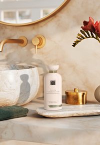 Witte, cilindervormige conditionerfles met groene accenten, geplaatst op een gemarmerde, crèmekleurige textuur. Bevat gouden hardware en een groene handdoek.
