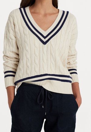 Vrouw draagt een crèmekleurige kabelgebreide trui met marineblauwe strepen op de V-hals, manchetten en zoom, gecombineerd met zwarte broek met trekkoord, handen in zakken.