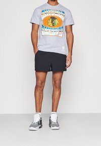 Hombre vistiendo una camiseta gráfica gris de los Chicago Blackhawks, pantalones cortos negros, calcetines blancos y zapatillas en blanco y negro, de pie contra un fondo sencillo.