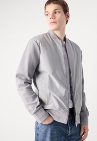 Jachetă bomber gri cu un față netedă și mâneci tricotate texturate. Include un închidere cu fermoar, manșete ribbate și buzunare laterale.