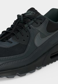 Μαύρο και σκούρο γκρι αθλητικό παπούτσι Nike Air Max με κλείδωμα κορδονιών, υφασμένα πάνελ και ορατή σόλα με αερόσακο σε ανοιχτό φόντο.