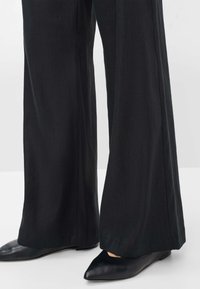 Pantaloni neri a gamba ampia realizzati in un tessuto morbido, caratterizzati da una silhouette pulita con una leggera lucentezza, abbinati a ballerine nere a punta.