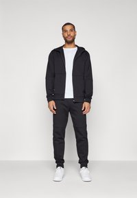 Svart zip-up hoodie med två framfickor, tillsammans med matchande svarta joggers. Vit t-shirt under och vita sneakers.