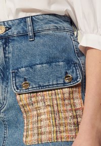 Shorts en denim avec une poche à patch ornée d'une texture tissée multicolore. Le matériel argenté sur la poche ajoute du détail au design.